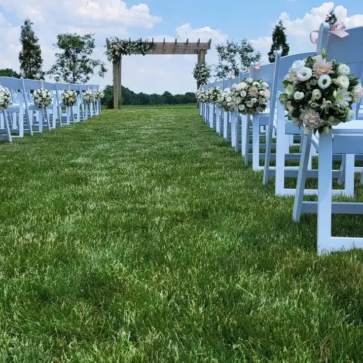 An outdoor wedding aisle lined with rows of white folding chairs, each decorated with floral bouquets, facing an arch.