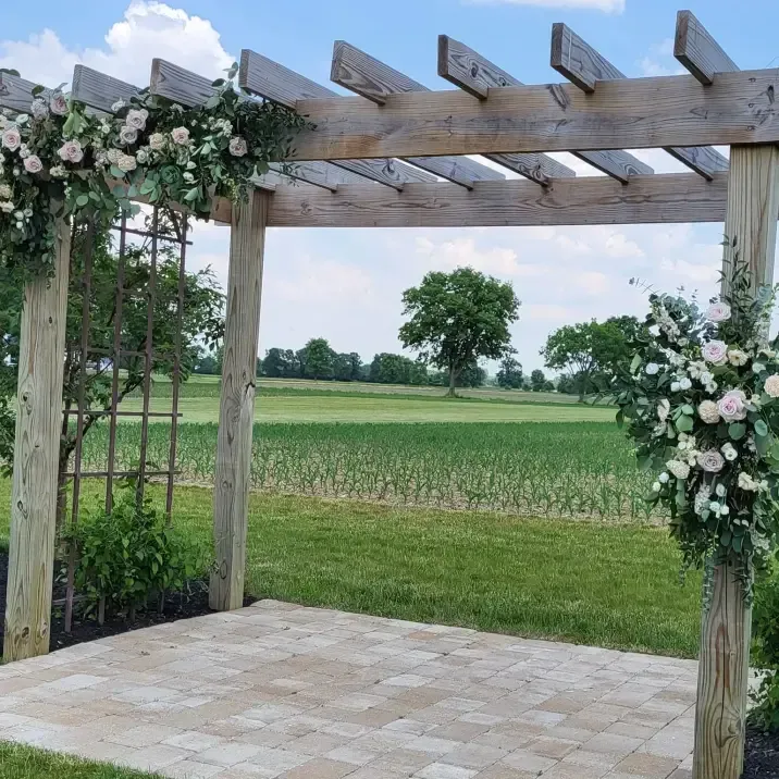 A wooden arbor decorated with white flowers stands on a stone patio overlooking a green field under a blue sky.