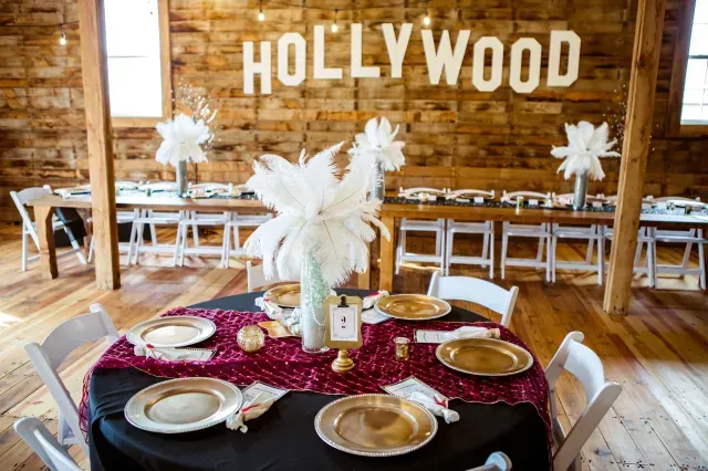 A round table set for a Hollywood-themed event with gold plates, a maroon runner, and feather centerpieces in a rustic barn.