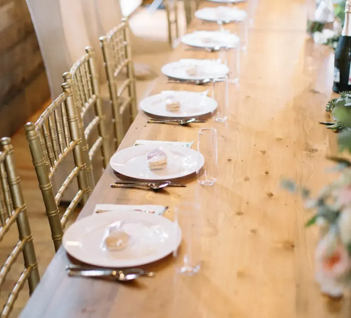A long wooden banquet table set for a formal event with white plates, gold chairs, and floral centerpieces.