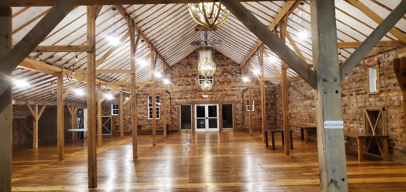 A rustic event hall featuring wood beams, high vaulted ceilings, stone walls, polished floors, and a hanging chandelier.