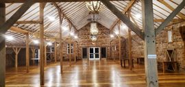 A rustic event hall featuring wood beams, high vaulted ceilings, stone walls, polished floors, and a hanging chandelier.