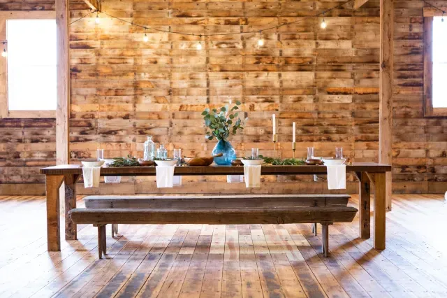 Rustic wooden table set with a blue vase and candles against a wood-paneled wall with string lights in a barn.