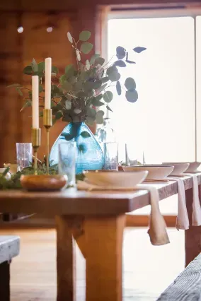A rustic wooden table set for dinner with a blue glass vase, eucalyptus stems, taper candles, and white place settings.