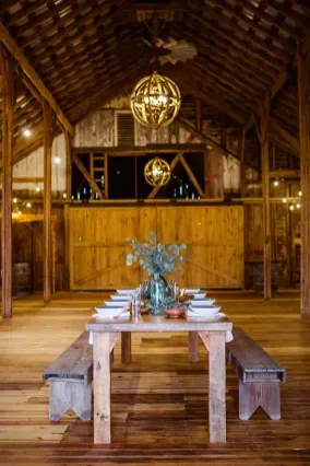 A rustic wooden barn interior features a long dining table with benches, set with white plates and a centerpiece.