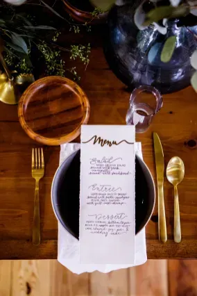 A place setting on a wooden table features a menu card inside a dark bowl, gold cutlery, and a glass against a dark vase.