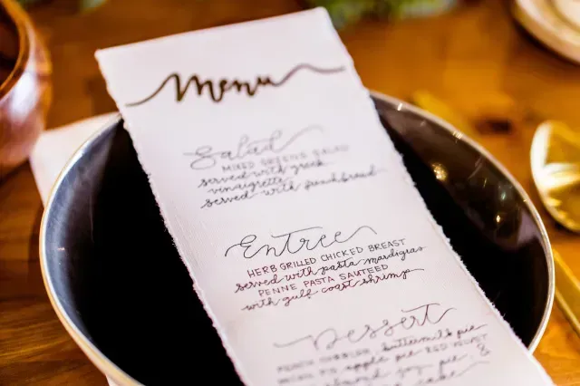 A handwritten menu on textured white paper sits inside a dark bowl on a wooden table, featuring salad, entree, and dessert.