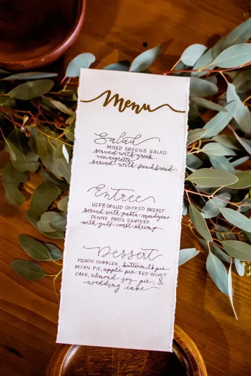 A handwritten wedding menu on torn white paper, placed on a wooden table among eucalyptus branches.