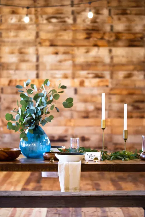 A rustic table setting featuring a blue vase with eucalyptus, candles, and a napkin against a wooden plank wall background.