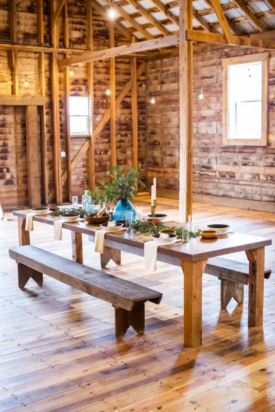 A rustic wooden table with a bench and place settings, decorated with blue vases and greenery in a wooden barn interior.
