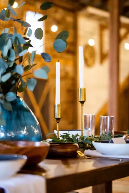 A table setting features a blue vase with greenery, two tall candles in brass holders, and bowls in a rustic barn venue.