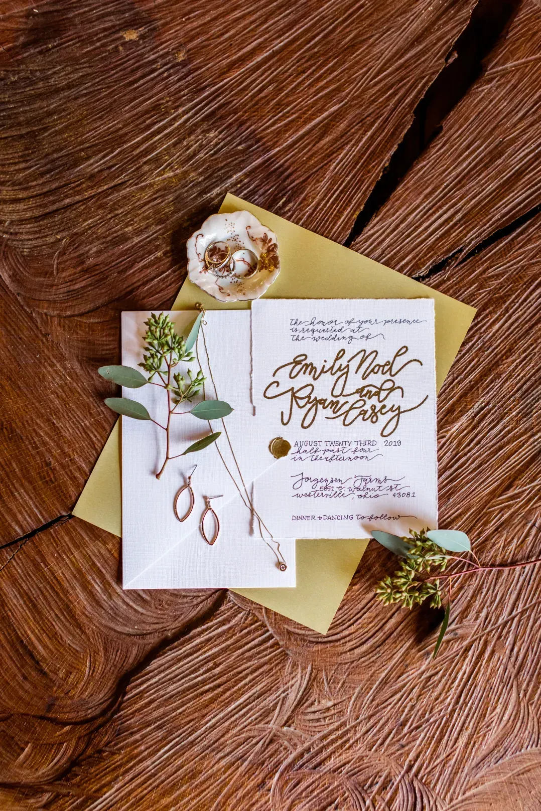 A wedding invitation, gold envelope, jewelry, and greenery arranged on a textured wooden background.