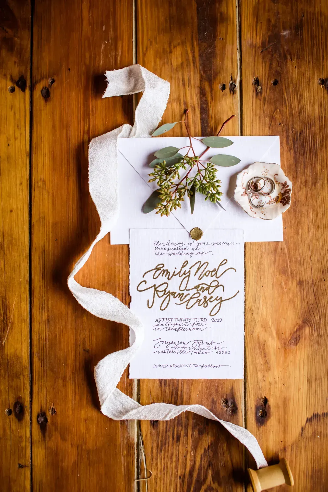 A wedding invitation, engagement rings in a dish, and greenery on a wooden surface with a long white silk ribbon.