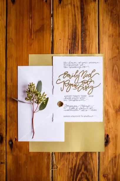 A wedding invitation with elegant gold calligraphy, adorned with a sprig of greenery, resting on a wooden surface.