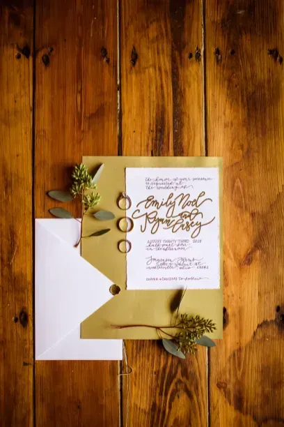 A wedding invitation with calligraphy sits on a gold cardstock backing, accented by rings and greenery on wood planks.