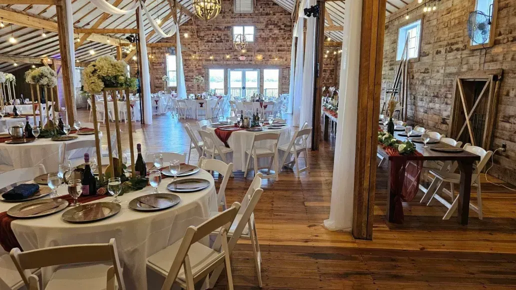 An indoor event venue with round dining tables, white tablecloths, wooden floorboards, and rustic stone walls.