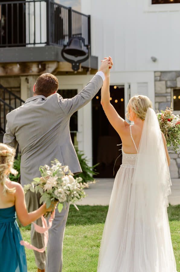 A wedding couple holding hands and walking toward a white building, with a person in a teal dress walking beside them.