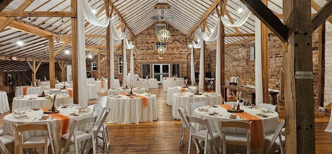A rustic barn interior prepared for a wedding with round tables, white tablecloths, orange runners, and hanging decor.