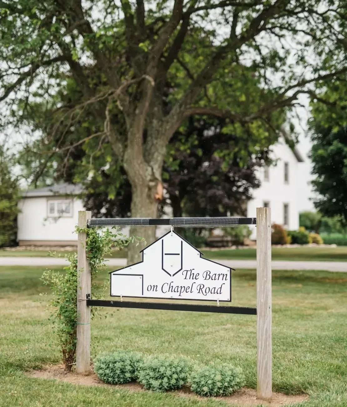 A white, barn-shaped sign hangs between two wooden posts on a green lawn, with a large tree and a house in the background.