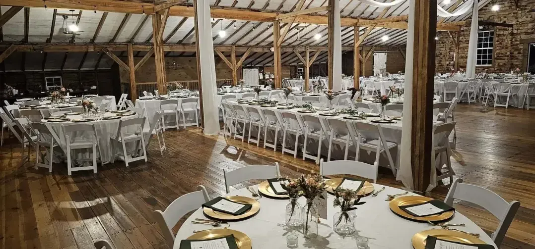 A rustic, wood-beamed wedding barn set with round and long tables covered in white linens, gold chargers, and chairs.