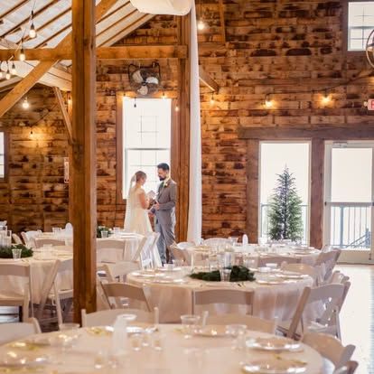 A bride and groom standing in an rustic barn venue surrounded by round reception tables with white tablecloths.