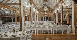 An indoor wedding venue inside a rustic barn with wooden beams, stone walls, white linens, and elegant drapery.