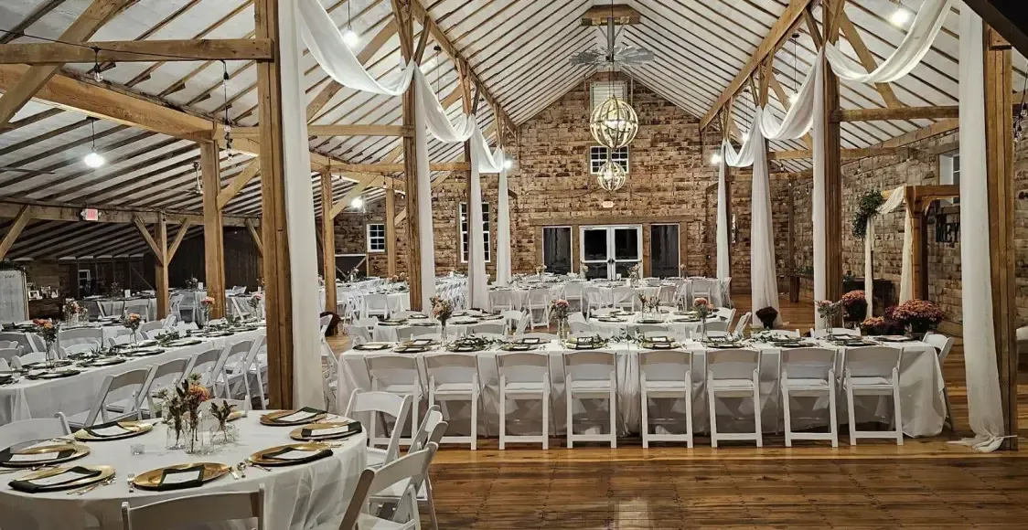 An elegant event venue inside a rustic timber-frame barn featuring round and long tables set for a formal dinner.
