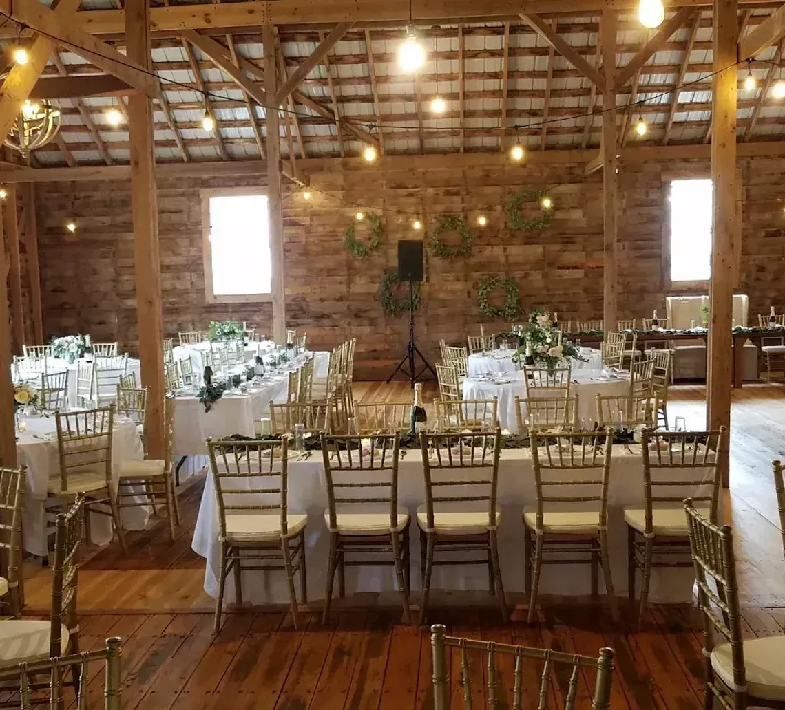 Rustic barn interior set for a wedding reception with round and long tables, gold chairs, and string lighting.