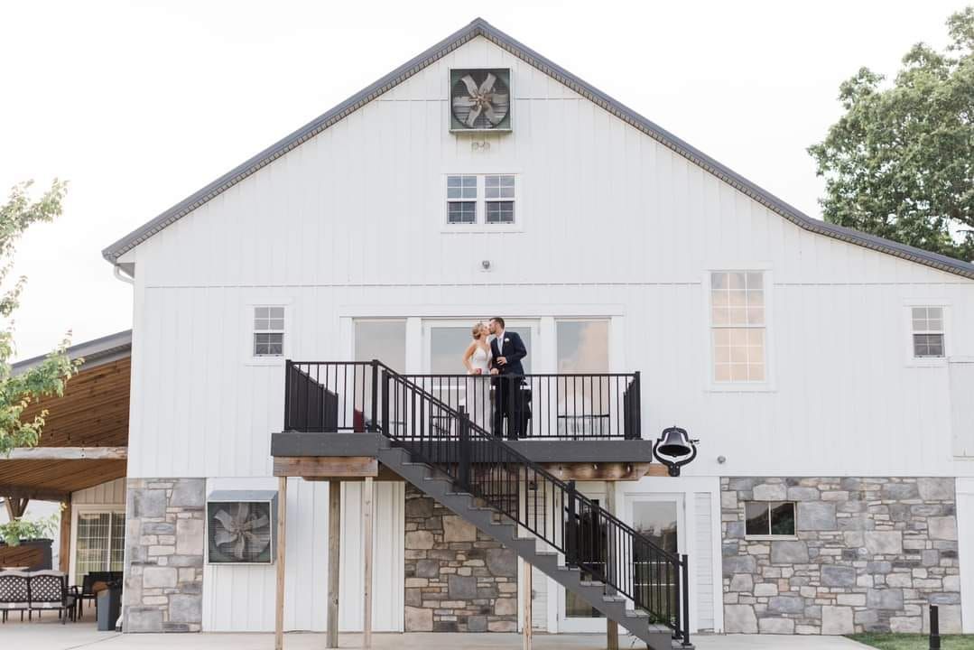 A bride and groom stand on the balcony of a white barn-style building with stone accents and an exterior staircase.