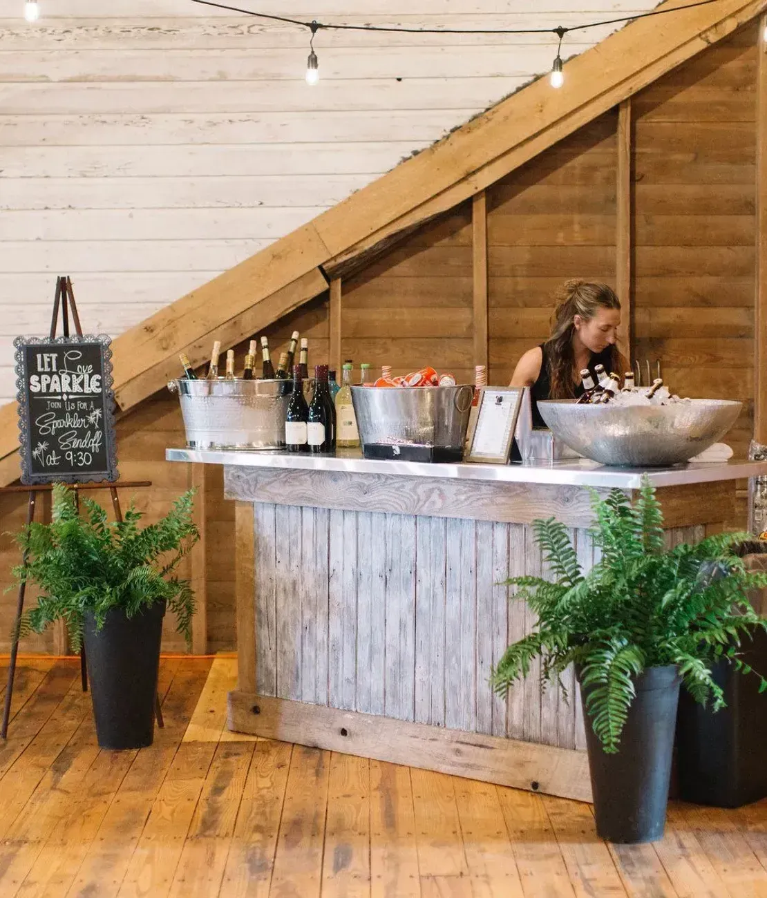 A bartender works behind a rustic wooden bar in a barn with string lights, plants, and a chalkboard menu.