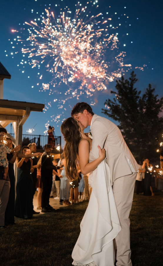 A couple in wedding attire kisses during an outdoor evening reception, with fireworks bursting in the sky above.
