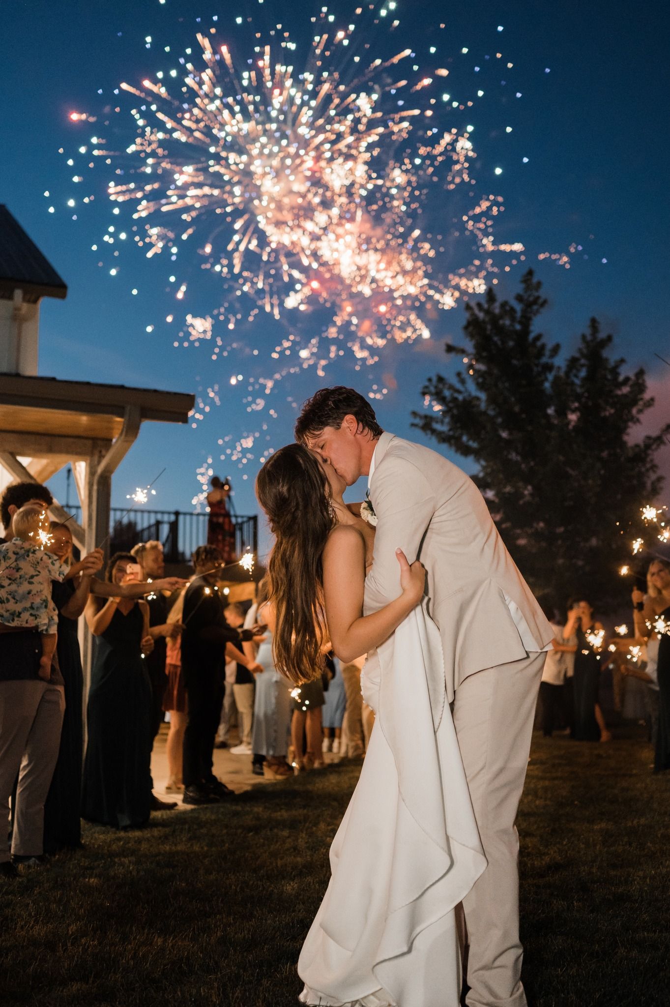 A couple kisses under a large fireworks display at night, surrounded by wedding guests in an outdoor venue.