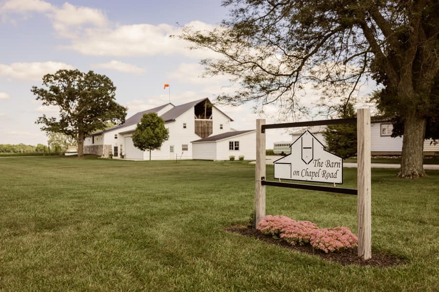 A white barn venue on a grassy lot with a wooden sign in the foreground reading 