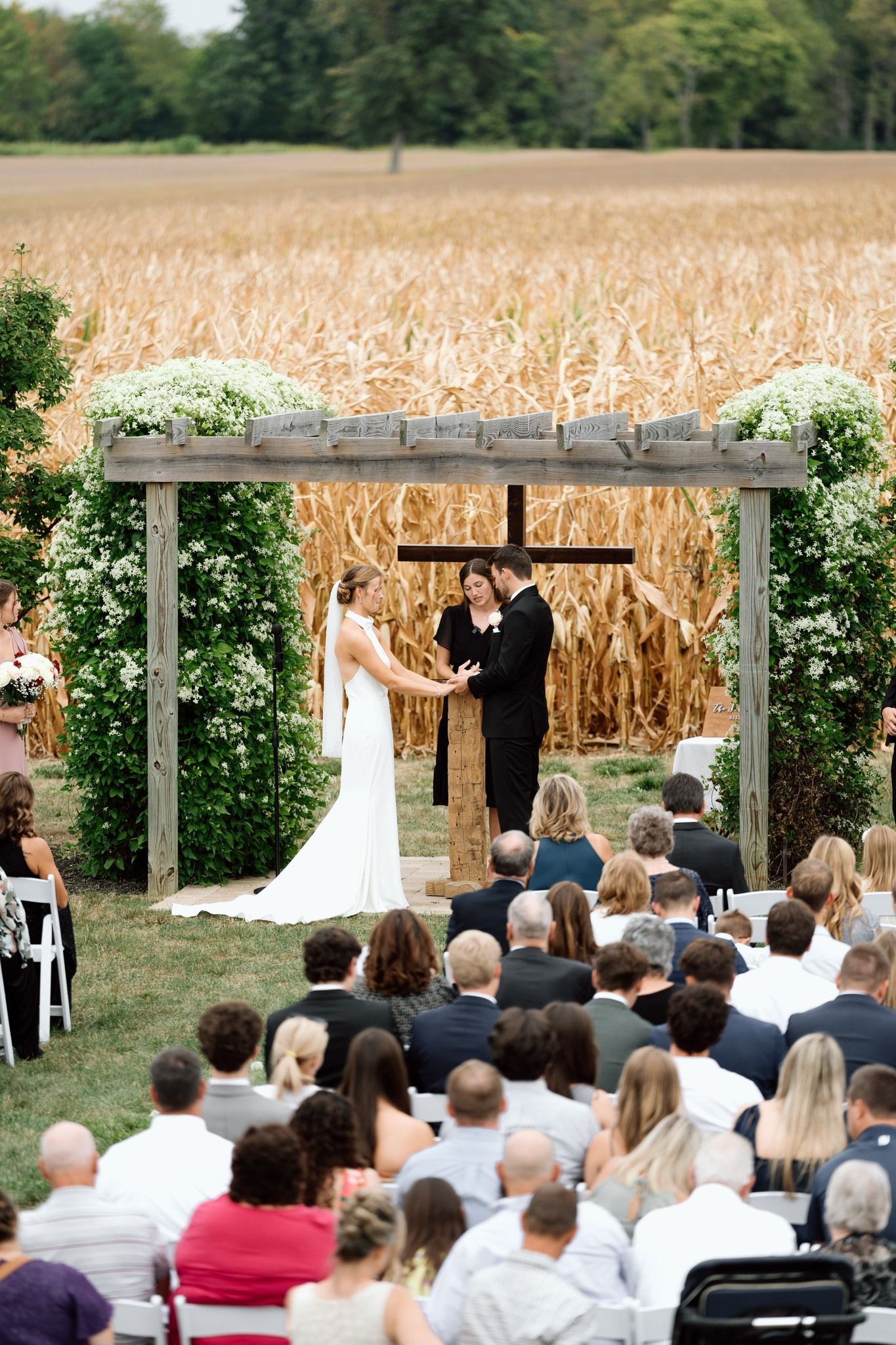 A wedding ceremony held outdoors in front of a rustic wooden arch and a large field of dry cornstalks.