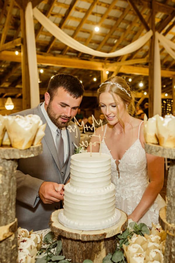 A couple in wedding attire cuts a tiered, white-frosted cake atop a rustic wooden stand in a dimly lit barn setting.