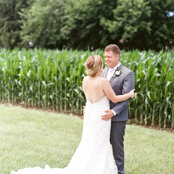 A bride in a white wedding gown and a groom in a grey suit embrace in front of a lush green cornfield.