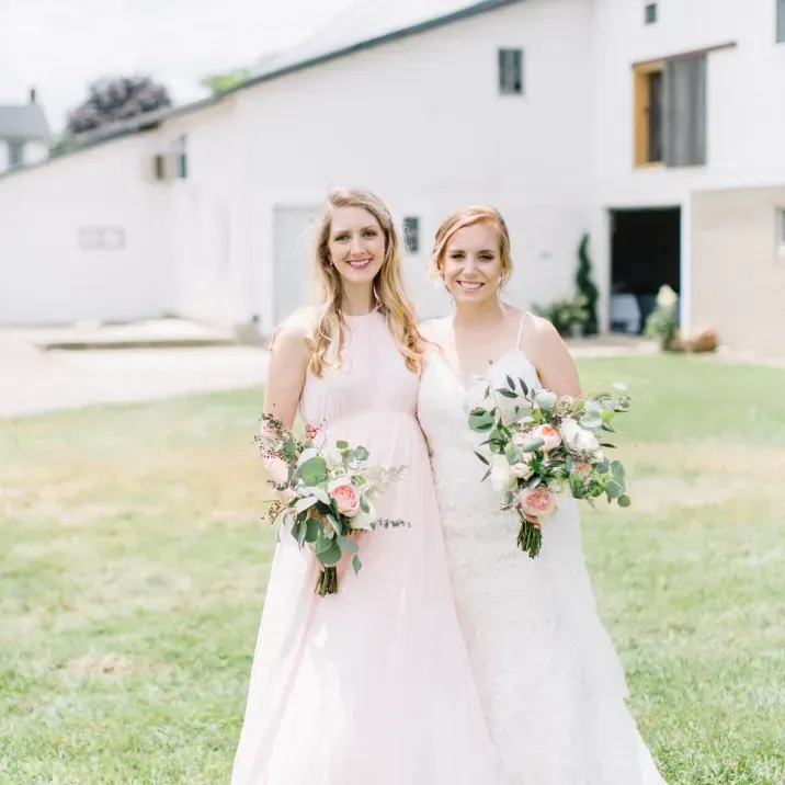 Two people in gowns hold bouquets while standing together on a lawn in front of a white building.