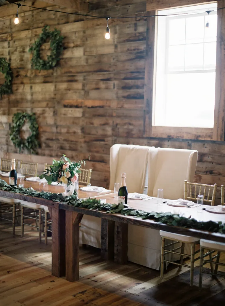A long, wooden wedding reception head table set with greenery, white linens, and tall, elegant chairs against a rustic wall.