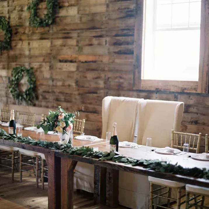 A rustic wooden table set for a wedding with greenery runners, champagne bottles, and high-backed white chairs.