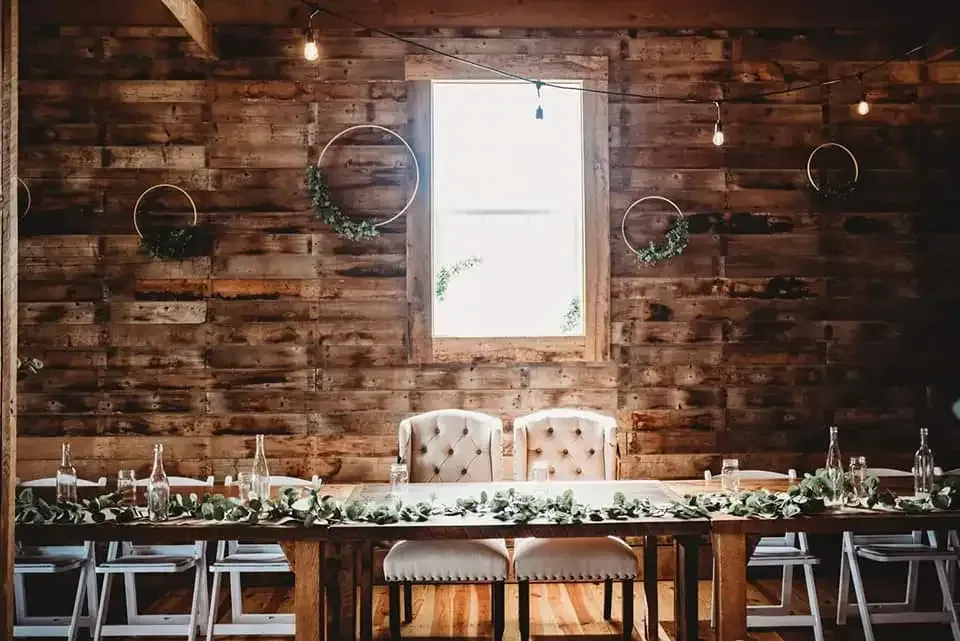 A rustic wooden wall features a centered window, decorative greenery hoops, and a long banquet table with two fancy chairs.