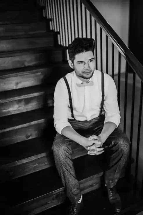 A person in a white button-down shirt, bow tie, and suspenders sitting on indoor wooden stairs, looking to the side.