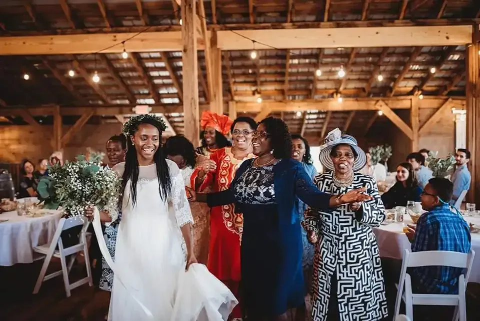 A bride in a white gown and flower crown leads a group of people dancing in a rustic, wood-beamed wedding reception hall.