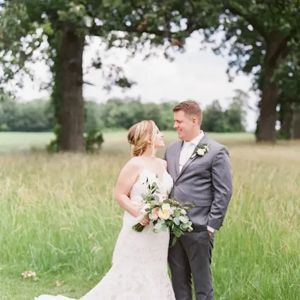 A couple in wedding attire stand holding a bouquet in a lush, grassy field with trees in the background.