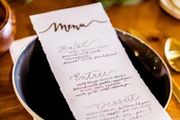 A handwritten menu rests on a dark bowl on a wooden table, detailing courses for salad, entree, and dessert.