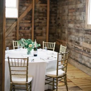 A round table with a white tablecloth, centerpiece, and gold chairs in a rustic room with wooden walls and flooring.