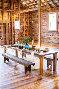A rustic wooden barn interior features a long dining table set with greenery, blue vases, and candles, flanked by benches.