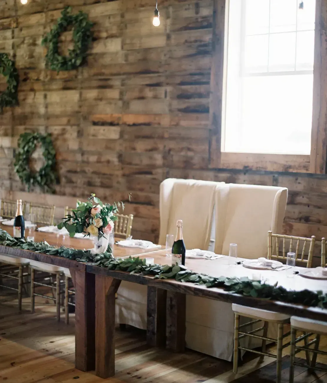 A long, wooden wedding reception head table set with greenery, white linens, and tall, elegant chairs against a rustic wall.