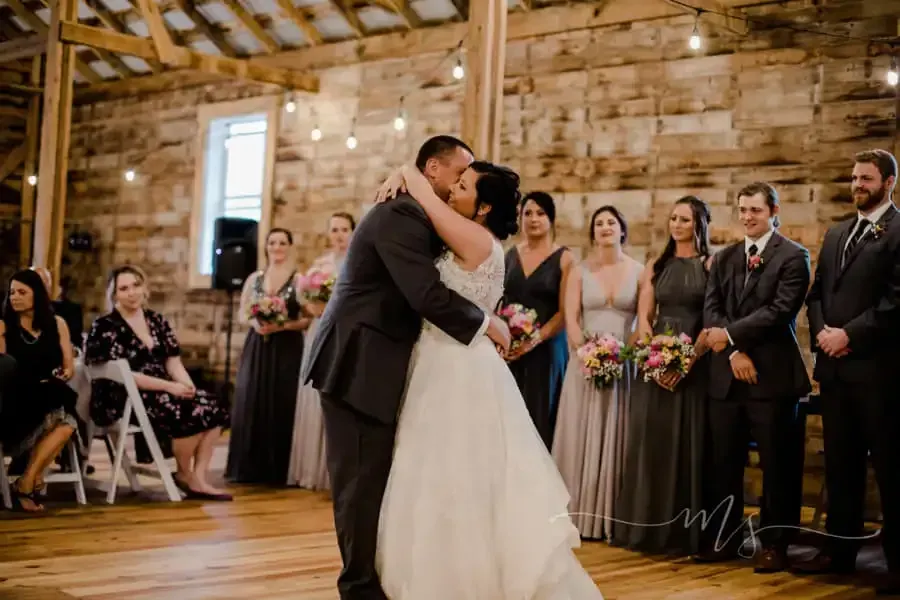 A bride and groom share a wedding kiss on a wooden dance floor in a rustic barn with wedding party members watching.