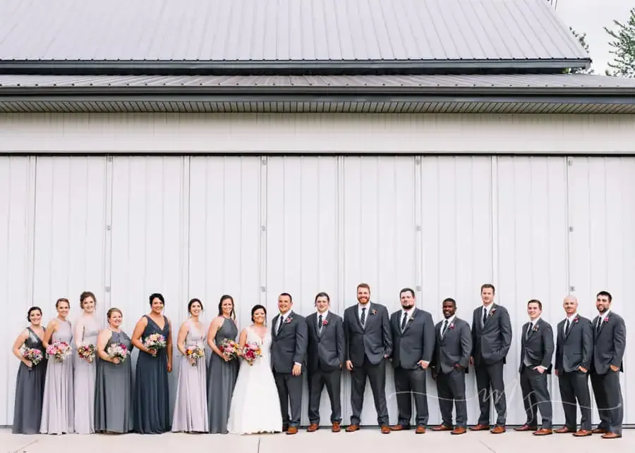 A bridal party stands in a line against a white barn wall. Everyone is dressed in formal wedding attire.