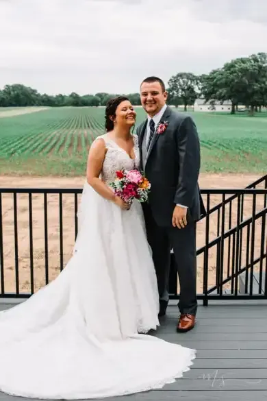 A bride and groom smile while standing on a deck overlooking a rural field.